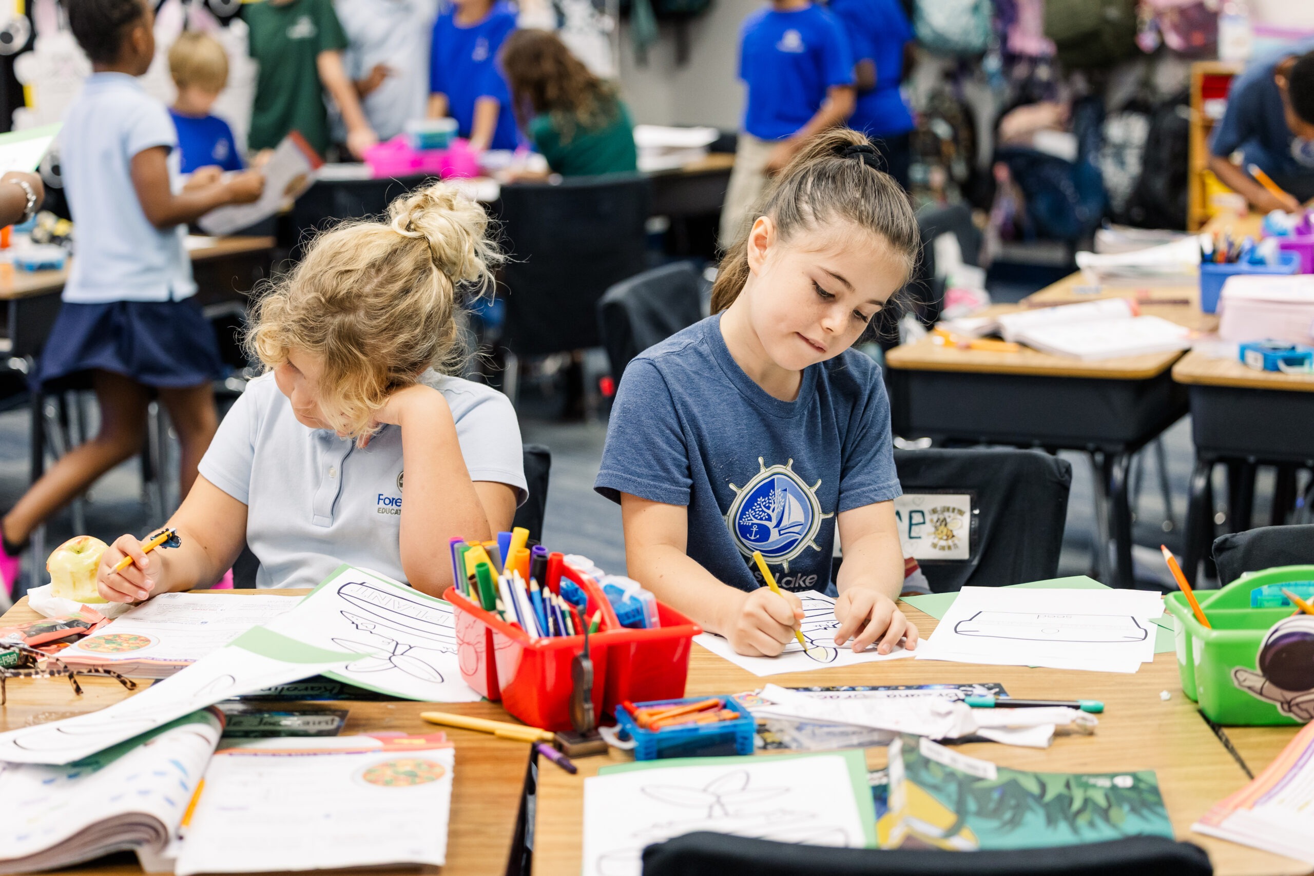Two Forest Lake Education Center students coloring worksheets together at a classroom desk with markers and colored pencils