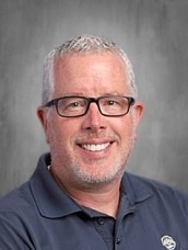 Smiling man with short gray hair and glasses wearing a navy polo shirt, posing for a portrait against a gray backdrop.