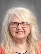 Portrait of an elderly woman with long white hair, wearing glasses and a coral blouse, smiling at the camera.
