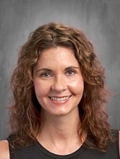 Portrait of a smiling woman with shoulder-length curly brown hair, wearing a dark sleeveless top, against a gray studio backdrop.