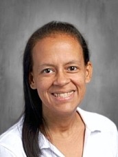 Portrait of a smiling woman with dark hair tied back, wearing a white blouse against a gray studio background.