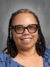 Smiling woman with braids, wearing dark glasses and a blue patterned blouse against a gray backdrop.