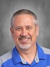 Smiling middle-aged man with short gray hair and beard, in a dark polo shirt, posing for a portrait against a blue background.