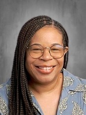 Smiling woman with long box braids, round glasses, wearing a blue patterned shirt in a neutral background.
