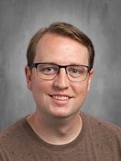 Head-and-shoulders portrait of a smiling man with short light brown hair and glasses, against a gray studio backdrop.