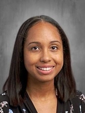 Portrait of a smiling woman with shoulder-length dark hair wearing a black floral blouse against a gray backdrop, a professional headshot for a profile.