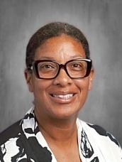 Professional headshot of a smiling woman with dark hair and black-framed glasses, wearing a black-and-white patterned blouse, against a gray studio backdrop.