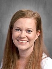 Portrait of a smiling woman with long red hair against a gray studio background, head and shoulders visible.