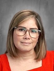 Portrait of a woman with shoulder-length light brown hair, wearing glasses and a red blouse, smiling slightly against a gray backdrop.