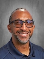 Smiling man with blue glasses wearing a navy polo shirt, posed against a gray studio backdrop.