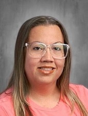Portrait of a woman with shoulder-length light brown hair, wearing clear-framed glasses and a pink shirt, smiling against a gray backdrop