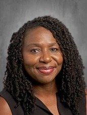 Portrait of a smiling African American woman with shoulder-length curly hair, wearing a dark sleeveless blouse, posed against a gray studio backdrop.