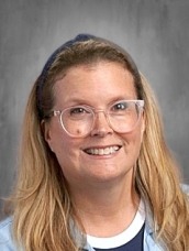 Smiling portrait of a blonde woman with glasses, wearing a blue cardigan over a white shirt, against a gray backdrop.