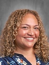 Portrait of a smiling woman with curly blonde hair wearing a navy floral blouse, against a gray backdrop.