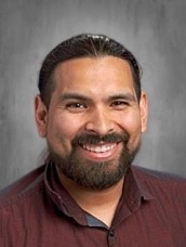 Portrait of a smiling man with dark hair and a beard, wearing a maroon button-down shirt, head-and-shoulders shot.