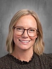 Smiling woman with blonde hair and glasses, wearing a dark patterned blouse, against a gray studio background.