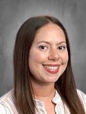 Portrait of a smiling woman with long brown hair in a white striped blouse against a gray studio backdrop to the right of center.