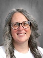 Portrait of a smiling woman with gray hair in two braids, wearing glasses and a white shirt against a neutral background.