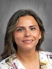 Head-and-shoulders portrait of a smiling woman with shoulder-length gray-blonde hair and a floral blouse against a gray backdrop.