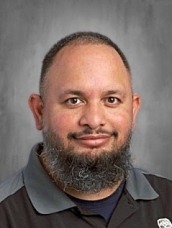 Head-and-shoulders portrait of a man with a gray beard wearing a dark polo shirt against a gray backdrop.