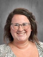 Smiling woman with shoulder-length wavy brown hair, glasses, and a light patterned blouse against a gray studio background.