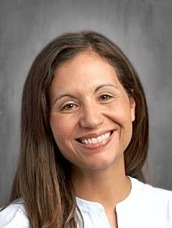 Smiling woman with long brown hair in a light blue blouse against a gray studio backdrop.
