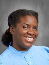 Smiling Black woman in a light blue blouse posing for a portrait against a gray studio background.