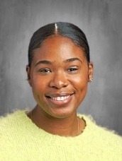 Portrait of a smiling woman with dark hair pulled back in a center part, wearing a lime-green sweater, against a gray studio backdrop.