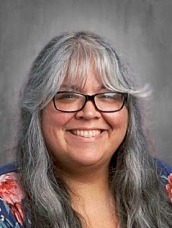 Portrait of a smiling woman with long gray hair and glasses, wearing a blue floral top against a neutral gray backdrop.