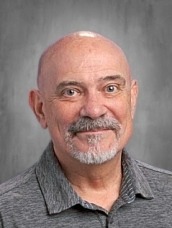 Portrait of an older man with a bald head and gray goatee, wearing a gray polo shirt and smiling at the camera.