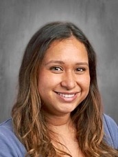 Smiling woman with long brown hair wearing a blue top, portrait against a gray backdrop.