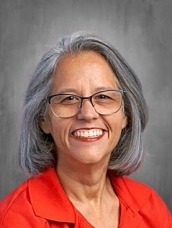 Portrait of a smiling woman with gray hair and glasses, wearing a red blouse, facing the camera.