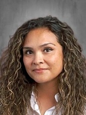 Professional headshot of a smiling woman with long, curly brown hair, wearing a black blazer and white blouse, against a gray studio backdrop.
