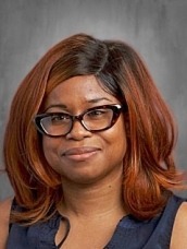 Professional headshot of a woman with auburn shoulder-length hair and black glasses, wearing a navy sleeveless top, smiling slightly.