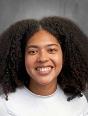 Portrait of a smiling woman with curly hair wearing a black sleeveless top, posing against a grey backdrop.