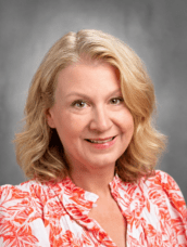 Smiling woman with shoulder-length blonde hair in an orange floral blouse against a gray studio backdrop.