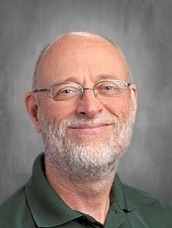 Portrait of a smiling older man with glasses and a gray beard, wearing a dark green polo against a neutral gray background.