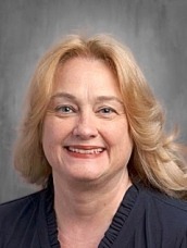 Professional headshot of a smiling blonde woman wearing a dark blazer against a neutral gray background.