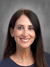 Portrait of a smiling woman with long dark hair, wearing a navy top, against a gray studio background.