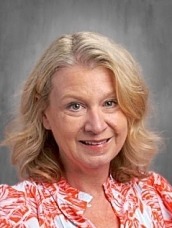Smiling woman with shoulder-length blonde hair in an orange floral blouse against a gray studio backdrop.