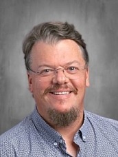 Portrait of a smiling man with glasses and a goatee, wearing a light blue patterned shirt, against a gray studio backdrop.