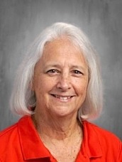 Smiling older woman with shoulder-length gray hair wearing a red polo shirt.”`,