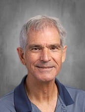 Smiling gray-haired man in a navy suit and light blue shirt, posing for a professional headshot against a neutral gray background.
