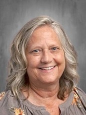 Smiling older woman with shoulder-length gray hair wearing a floral blouse, against a gray studio backdrop.