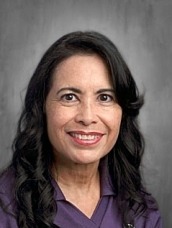 Professional headshot: smiling woman with dark hair in a purple blouse, gray studio backdrop.