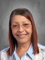 Head-and-shoulders portrait of a smiling woman with brown hair and glasses, wearing a light blue blouse and lace cardigan against a gray background.