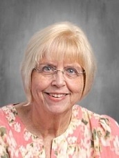 Portrait of a smiling elderly woman with short blonde hair, wearing glasses and a pink floral blouse.
