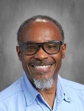 Smiling man with glasses, short gray beard, wearing a light blue shirt, against a gray studio backdrop.
