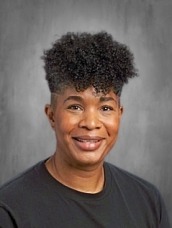 Smiling person with short curly hair wearing a black shirt against a gray studio backdrop.