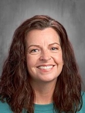 Portrait of a smiling woman with shoulder-length wavy brown hair in a teal top, against a gray studio backdrop.